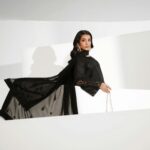 A poised model in a black eastern dress poses indoors, showcasing elegance and style.